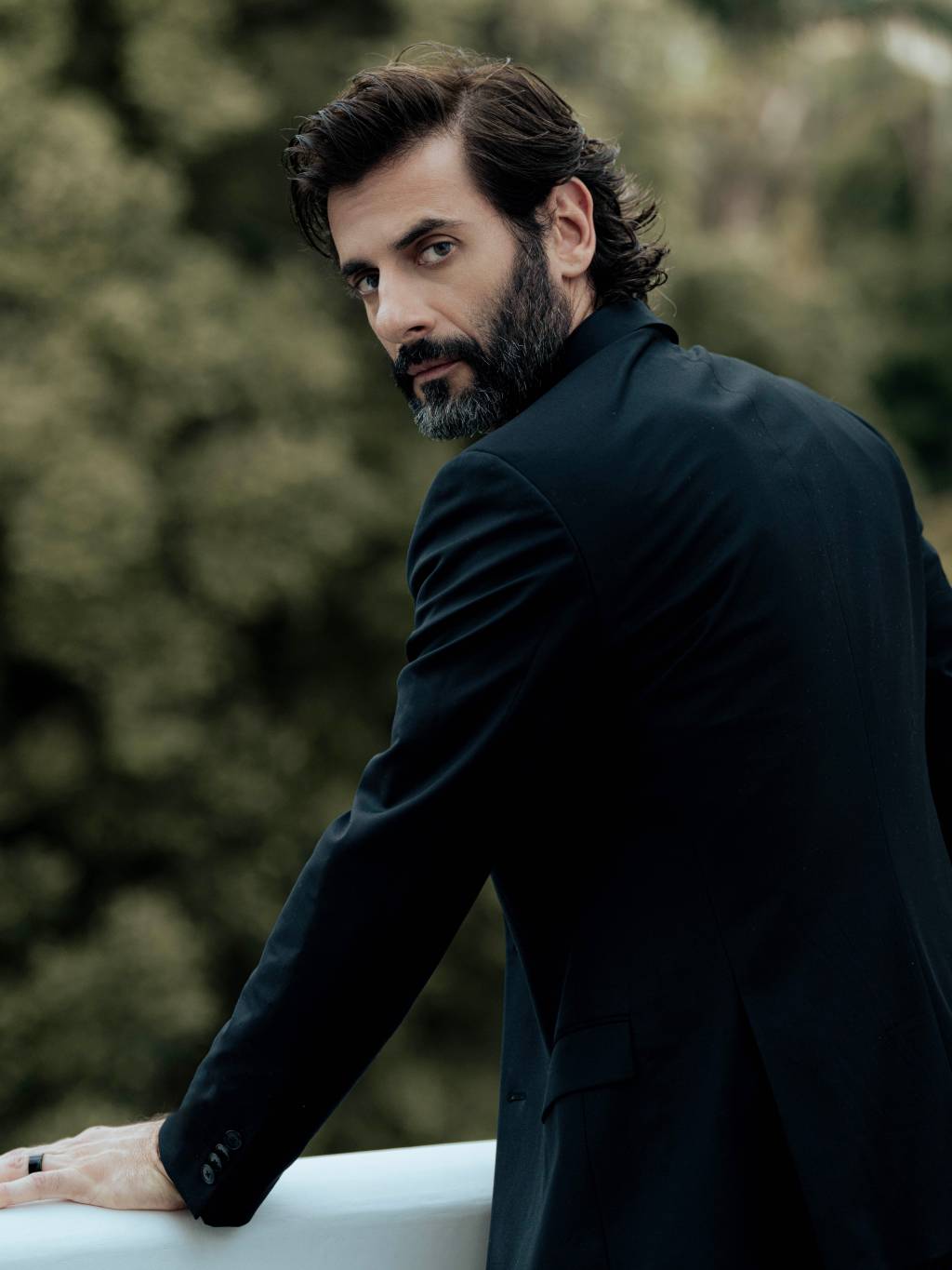 Flávio Tolezani (Vinícius Mochizuki-Divulgação) (1) Homem branco de barba e cabelo pretos posa apoiado em sacada, voltado de costas, olhando para a câmera. Veste paletó preto.