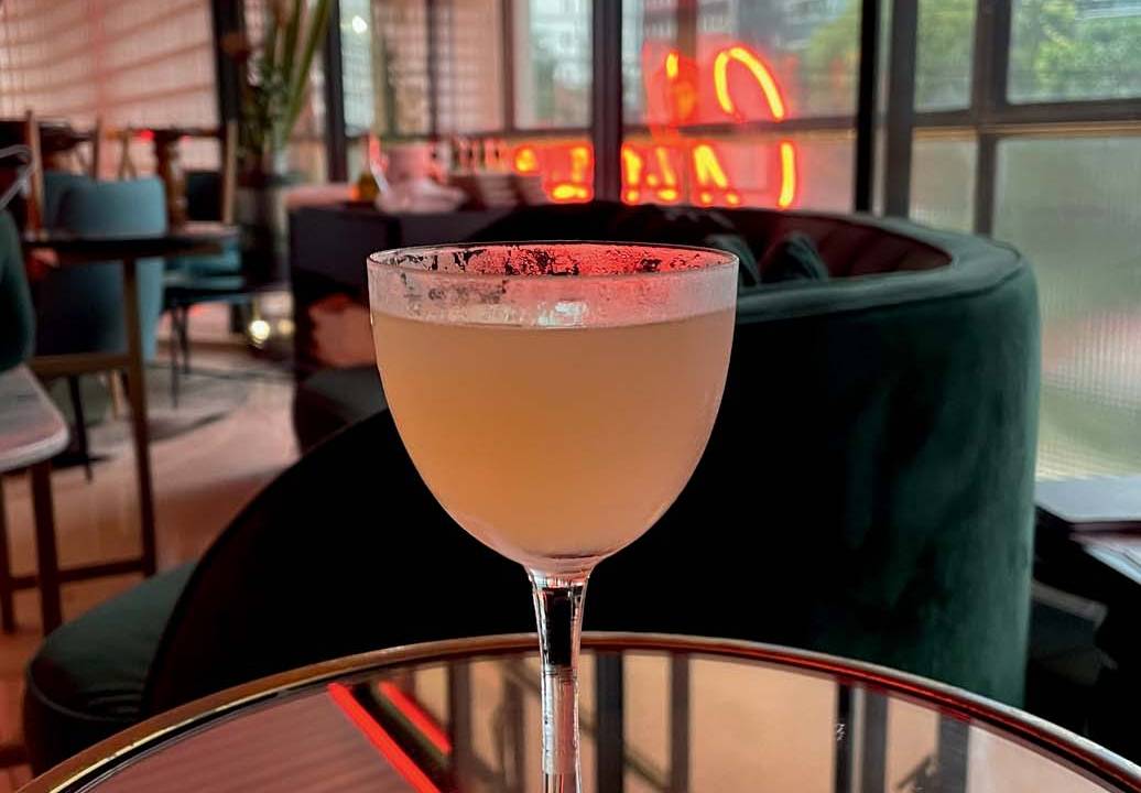 Drinque em taça coupe em tom laranja claro sobre mesa redonda espelhada com poltronas verdes ao fundo