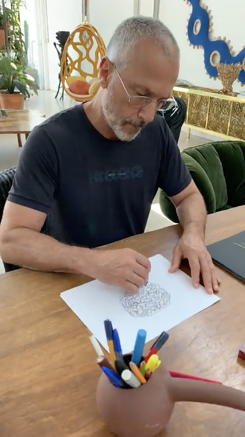 Humberto_Drawing_Bacana_April_2023 Homem grisalho sentado em mesa de madeira faz rascunho em papel. Veste camiseta escura.