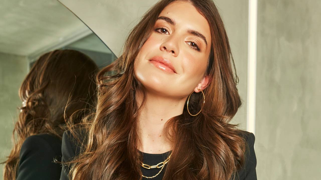 Manuela Bordasch posa apoiada em espelho. tem cabelos longos castanhos e veste blazer e camiseta preta.