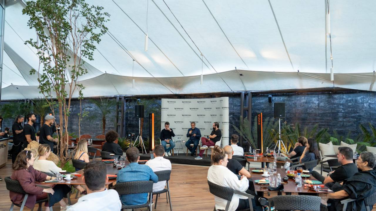 Mesas em restaurante virado para painel com três pessoas debatendo.