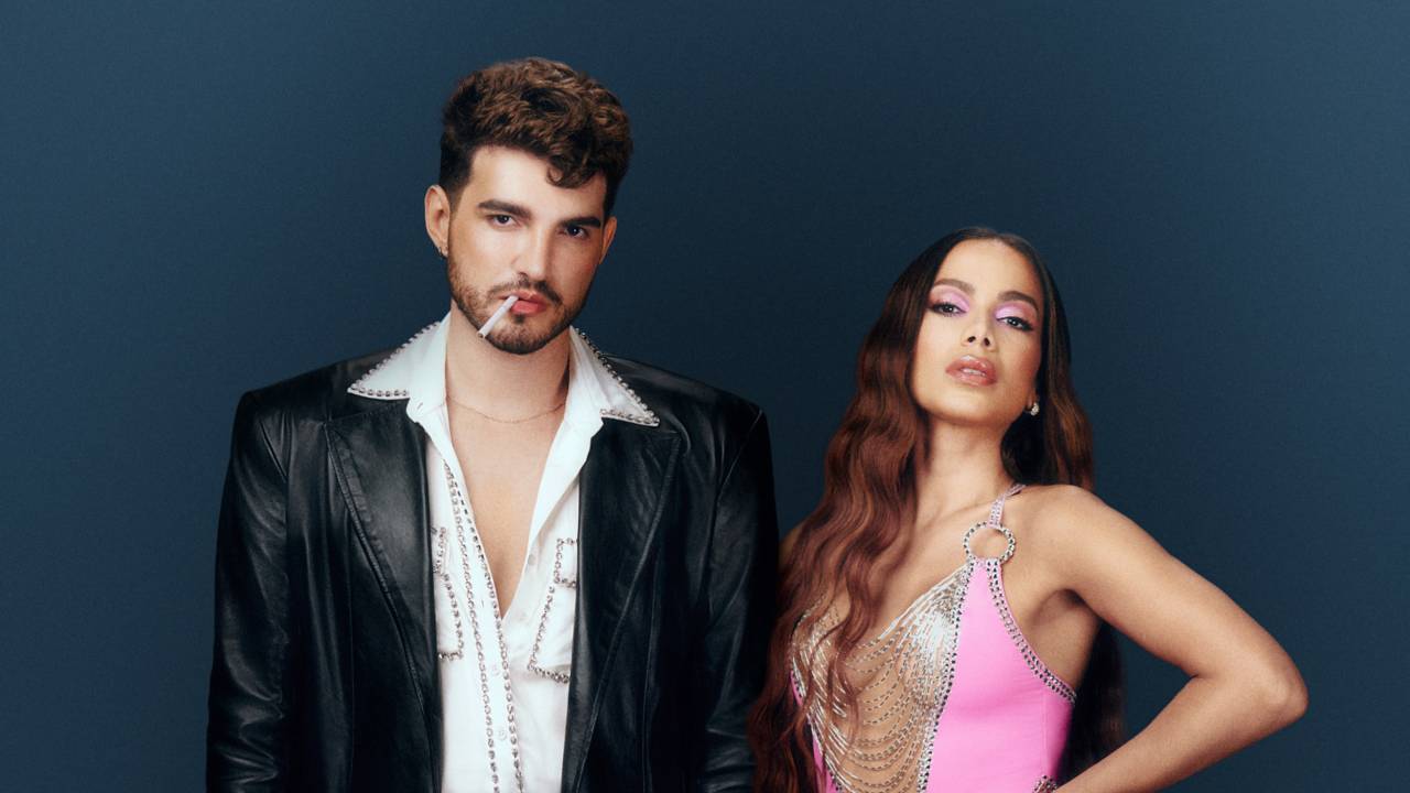 Jão e Anitta posam de pé lado a lado. Ele veste terno preto e camisa branca com peito aberto, ela veste vestido decotado rosa com parte em formato de V na cor vermelha.