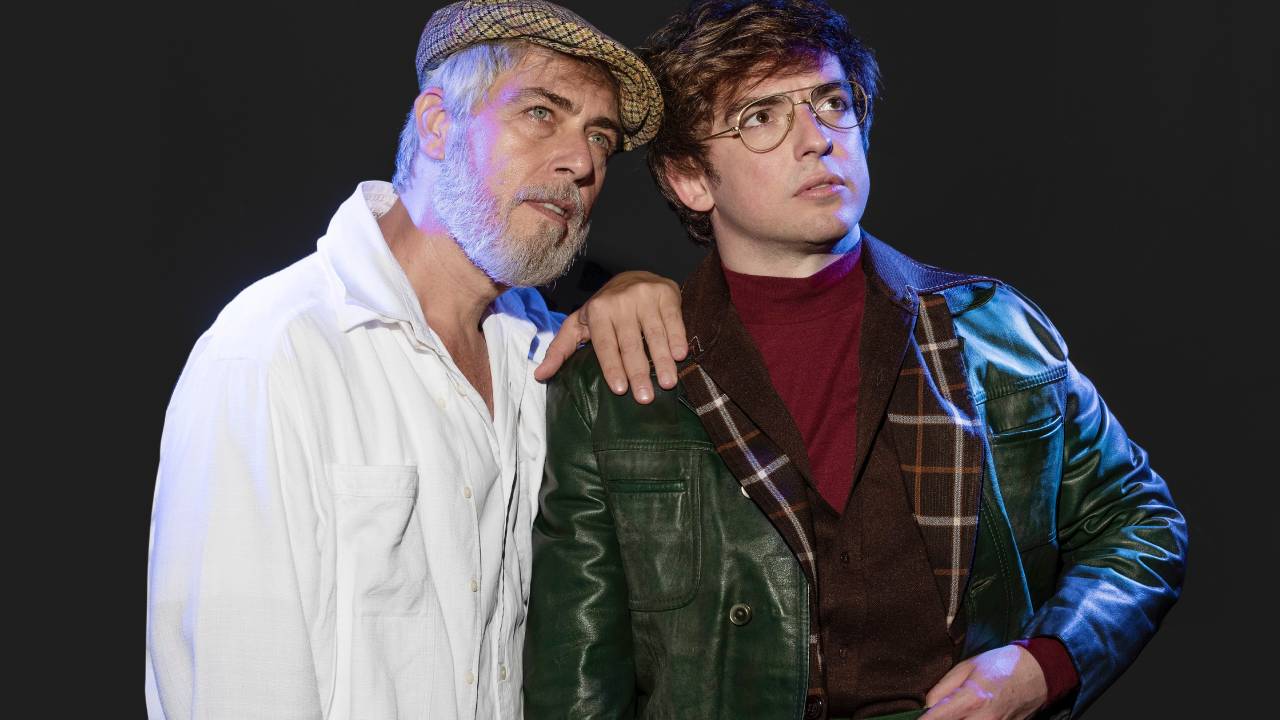 Dois homens posam olhando à direita. O da esquerda é grisalho e veste camisa branca. O da direita, mais jovem, veste jaqueta preta e camiseta cor de vinho.