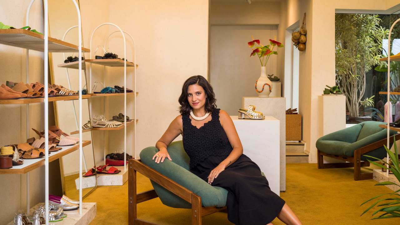 Mulher de vestido preto posa apoiada em poltrna verde e de madeira com prateleiras de sapatos à esquerda.