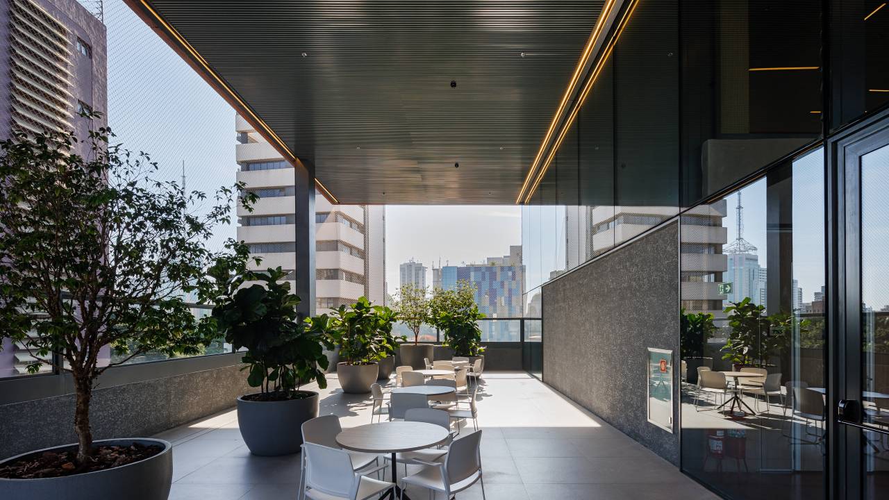 Imagem exibe terraço iluminado, com plantas e algumas mesas e cadeiras brancas.