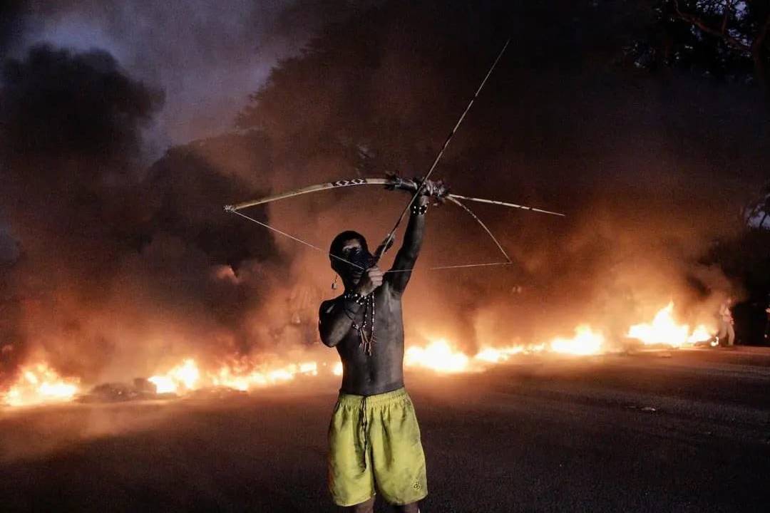 Indígena com arco e flecha apontados para o céu. Atrás, um grande fogaréu.