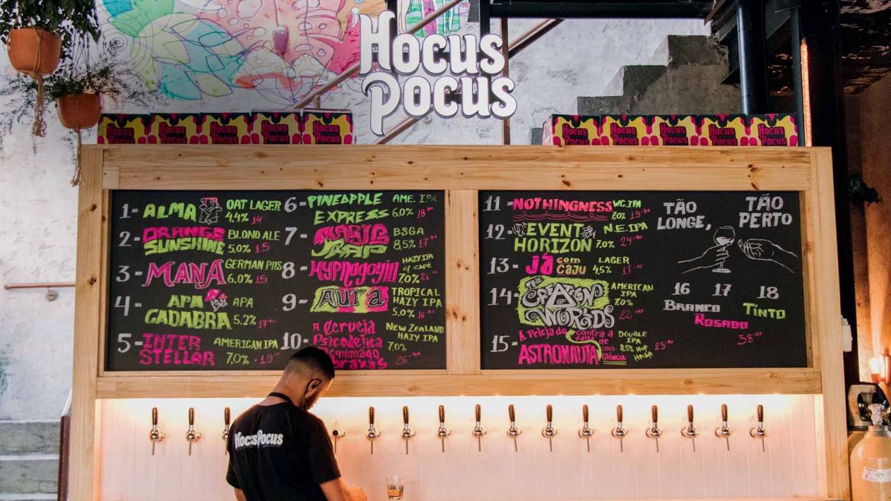 Parede no bar da Hocus Pocus com lousa descrevendo chopes e torneiras com homem servindo copo abaixo.