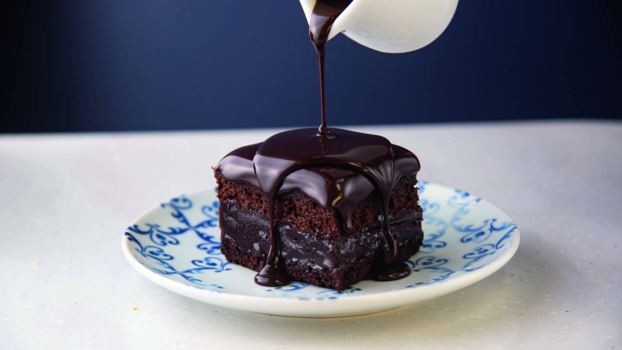 Prato de louça branca e azul com pedaço quadrado de bolo de chocolate com calda sendo derrubada por cima em mini jarra.