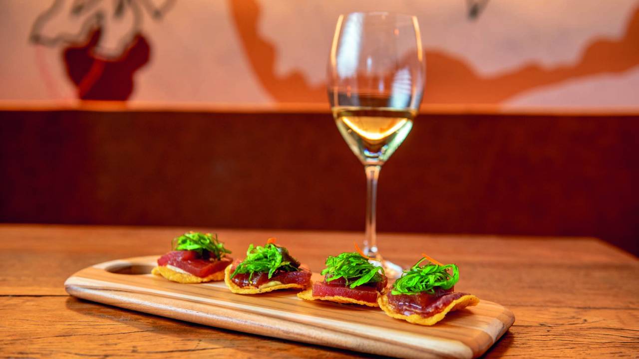 Tábua de madeira sobre mesa com quatro canapés de atum ao lado de taça de vinho branco.