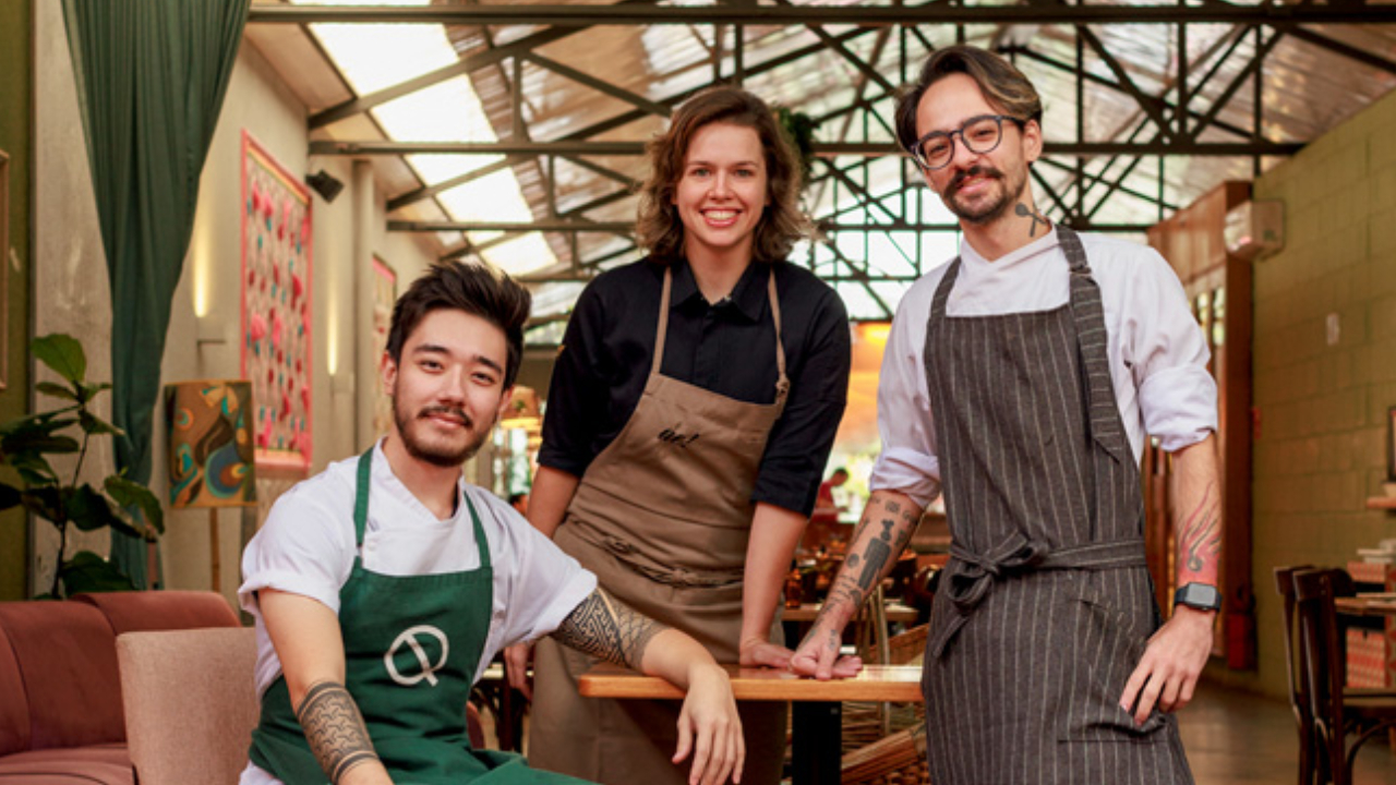 Três chefs de cozinha em salão de restaurante. Sentado à esquerda, homem asiático, ao centro mulher branca apoiada em mesa e à direita homem em pé.