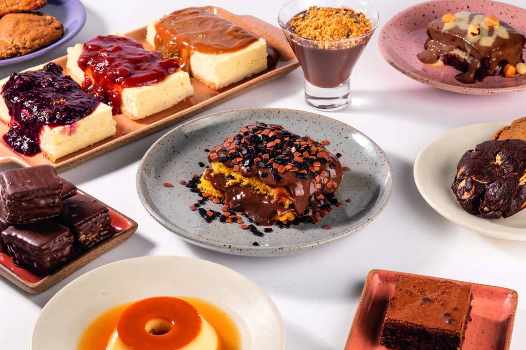 Papila Deli Diferentes tipos de sobremesa (bolo de cenoura, cheesecake, pudim, mousse) sobre superfície branca.