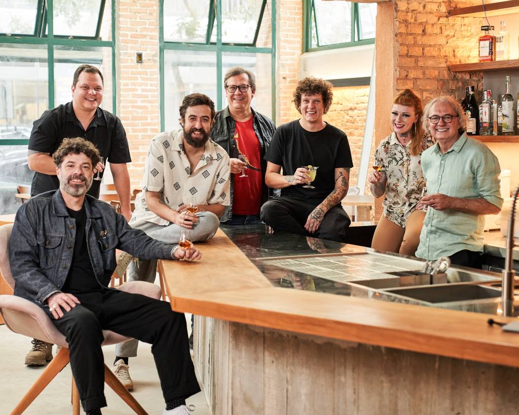 Bar dos Cravos Seis homens brancos e uma mulher branca sentados no balcão de um bar olhando para a foto.