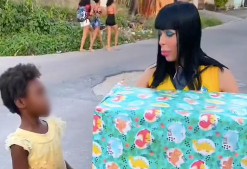 Uma mulher branca de cabelos pretos e lisos com uma grande caixa de presente nas mãos olhando para uma criança negra