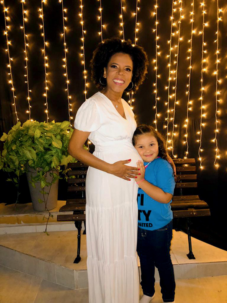 luciana1.jpeg Mulher negra posa sorrindo ao lado da filha. Está grávida e veste um vestido inteiro branco. Ao fundo, luzes em cordas preenchem toda a parede.