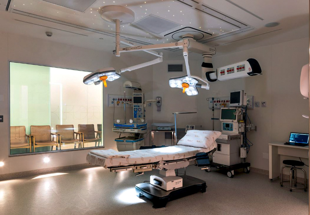 vista pro quarto em baixa.JPG Imagem de quarto hospitalar com grande janela, maca e equipamentos no teto.