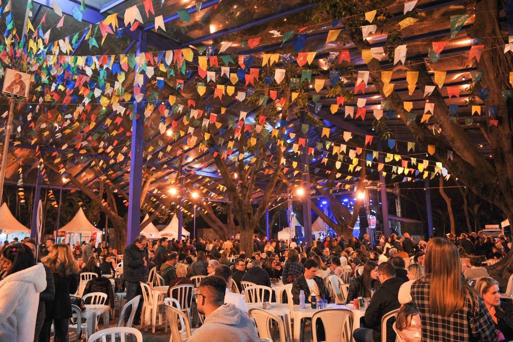 11 – Esperia (Ustreming Creative Content-Click Eventos-Divulgação) Mesas e cadeiras de plástico branco são ocupadas por pessoas e no topo aparecem fileiras de bandeirinhas coloridas.