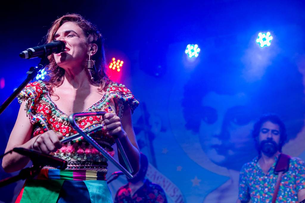 2 – Marina Aydar (Divulgação) Cantora de cabelos castanhos lisos fala ao microfone de olhos fechados, em palco com luz e fundo azuis.