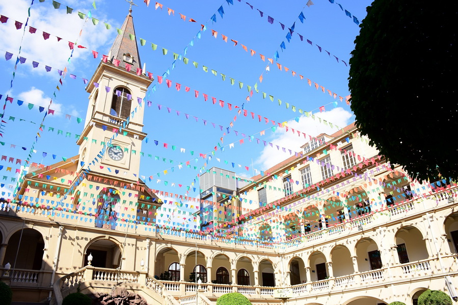 4 – Colégio Marista Arquidiocesano (Arquivo Pessoal) (1) Pátio de colégio com bandeiras coloridas cruzadas no céu. O local tem uma torre com telhado pontudo.