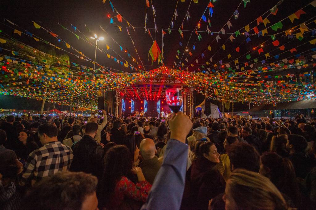 7 – Paineiras (Clube Paineiras do Morumby-Divulgação) Homem ergue taça de vinho no primeiro plano, com multidão à frente de olho em um palco iluminado. No topo, fileiras de bandeirinhas coloridas.