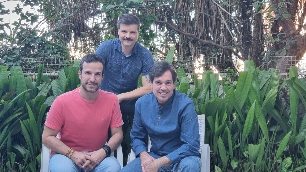 Três homens brancos posam sentados em área verde. Um veste camiseta vermelha, os outros dois camisa azul. O do meio está apoiado atrás dos dois.