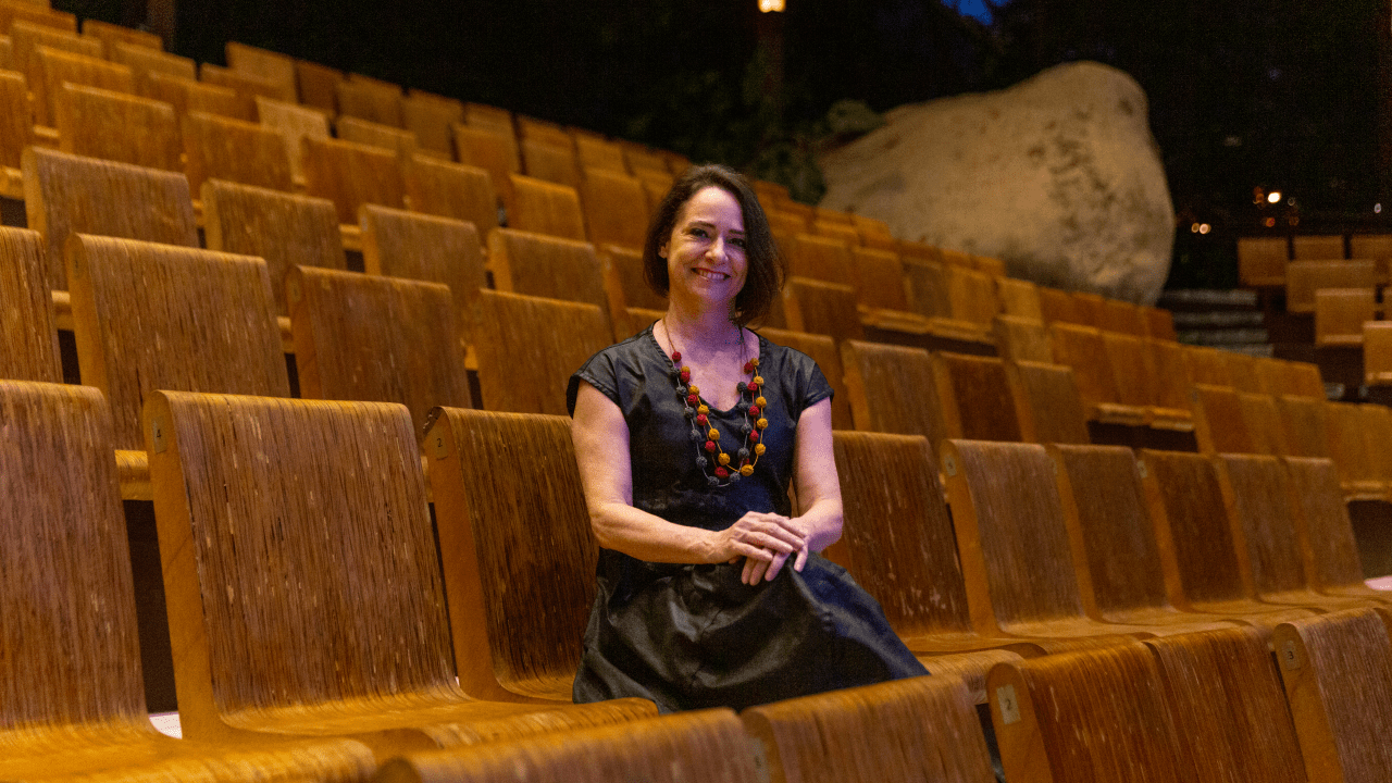 Mulher branca de cabelos pretos curtos aparece sentada de pernas cruzadas em uma das poltronas marrons em fileiras de teatro.