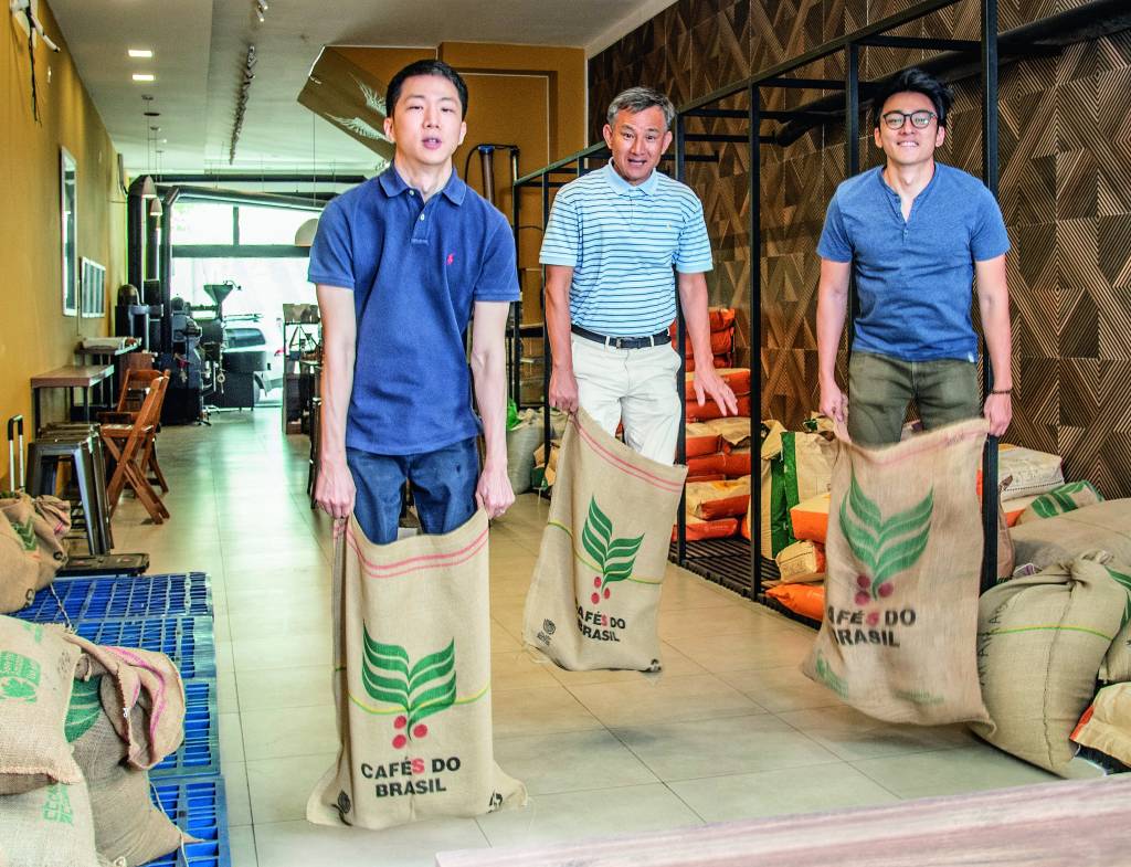 Um Coffee Co. Três homens asiáticos fazendo corrida do saco com sacos de café dentro de uma cafeteria.