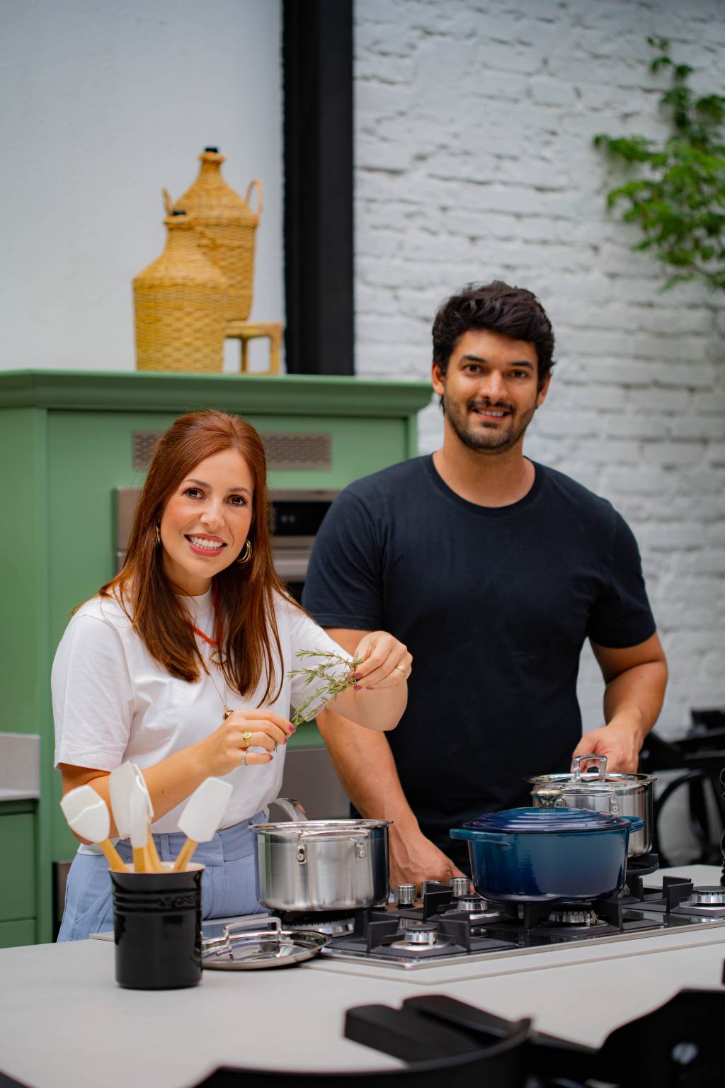 Ana Lembo e José Soares (Matheus Viotti-Divulgação) Mulher branca e ruiva posa ao lado de homem com cabelos e barba pretos. Ela veste camisa branca e ele uma camiseta preta. Ambos estão em uma cozinha, manipulando utensílios de culinária.