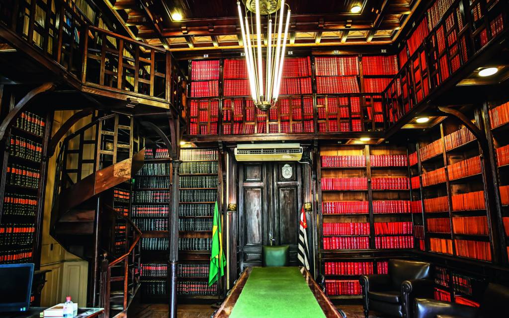 BIBLIOTECA_SECJUSTICA_1.JPG Biblioteca com piso verde, detalhes vermelhos e prateleiras pretas. Um grande lustre aparece no centro.