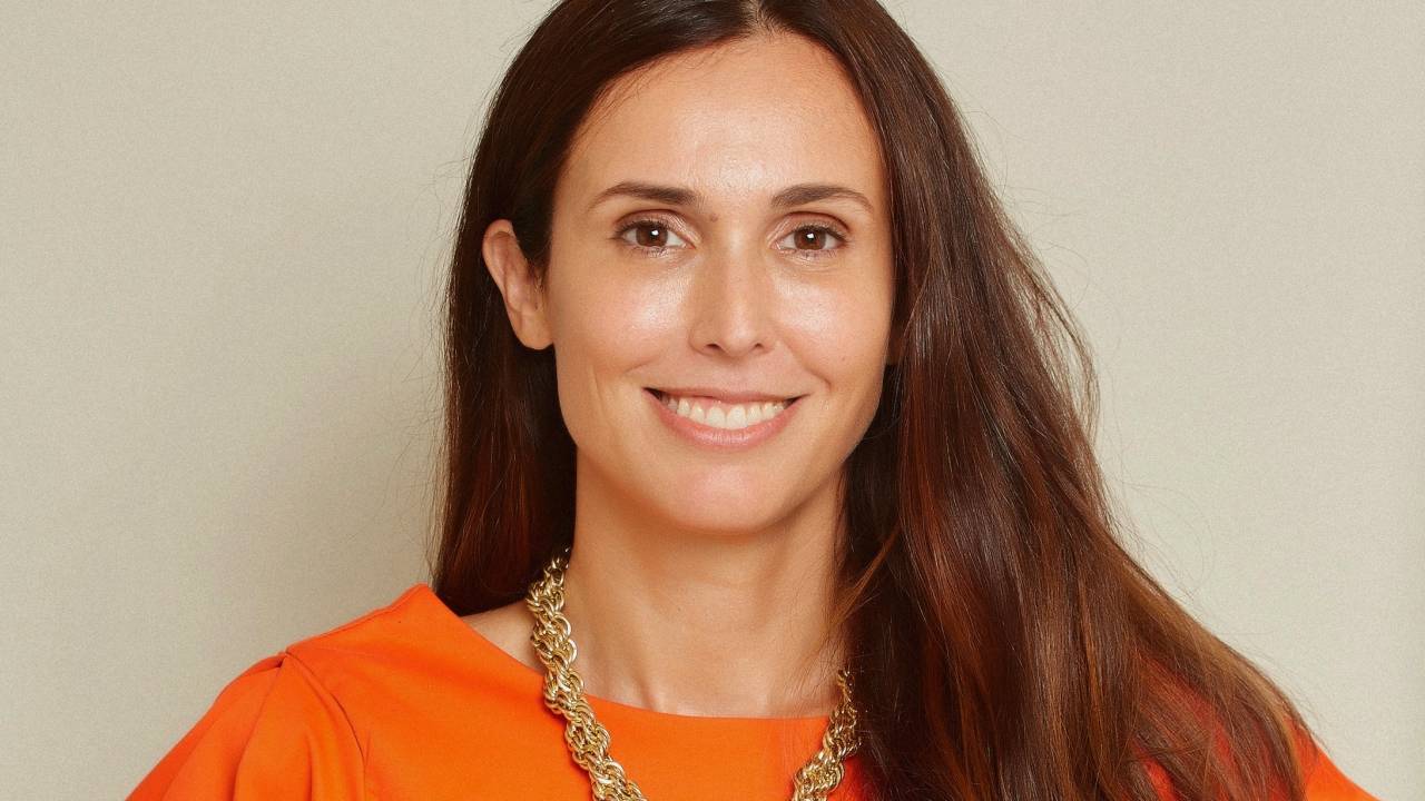 Moça branca de cabelos lisos castanhos sorri com uma mão na cintura. Veste blusa laranja e calça amarela, com colar dourado.