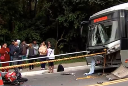 Imagem mostra ônibus com frente danificada e moto destruída, entre outros destroços em uma pista de asfalto. Ao fundo, pessoas estão reunidas próximas ao local do acidente