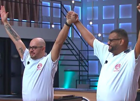 Imagem mostra Diego à esquerda e Wilson à direita, ambos de braços levantados e mãos dadas. Eles estão no cenário do Masterchef e vestem roupas de chefs de cozinha