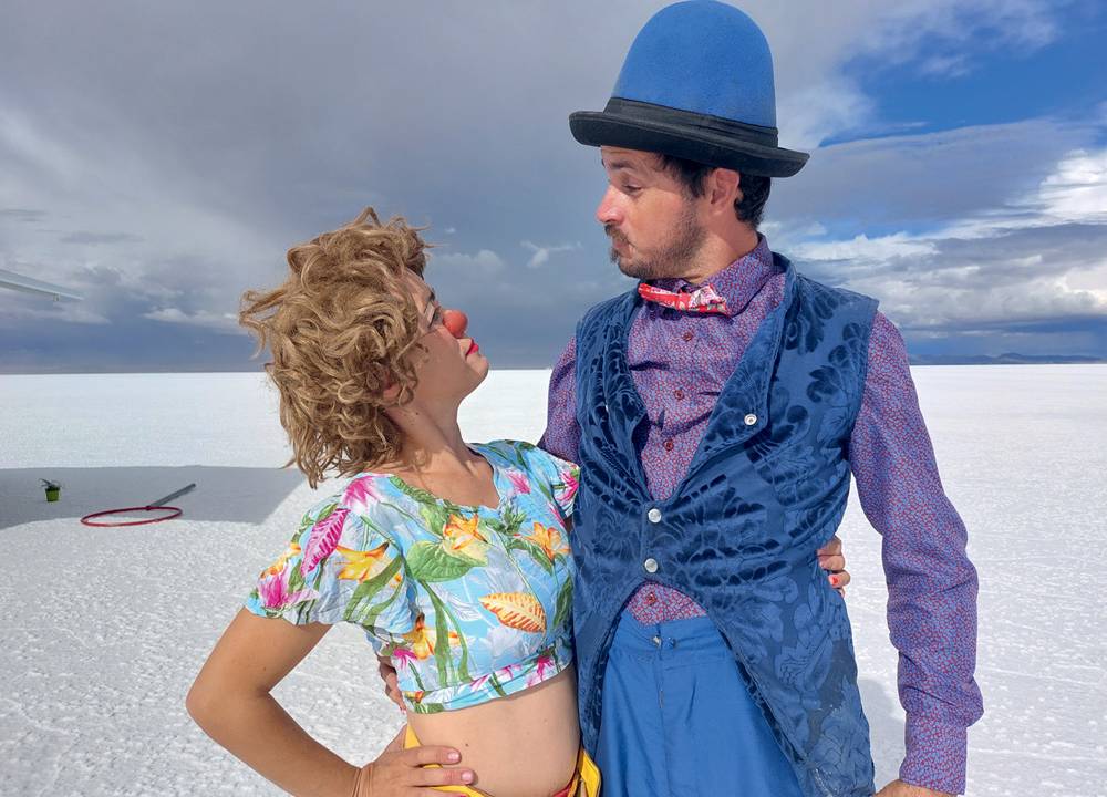 Casal com roupas de circo se abraçam e se encaram em cenário a céu aberto.