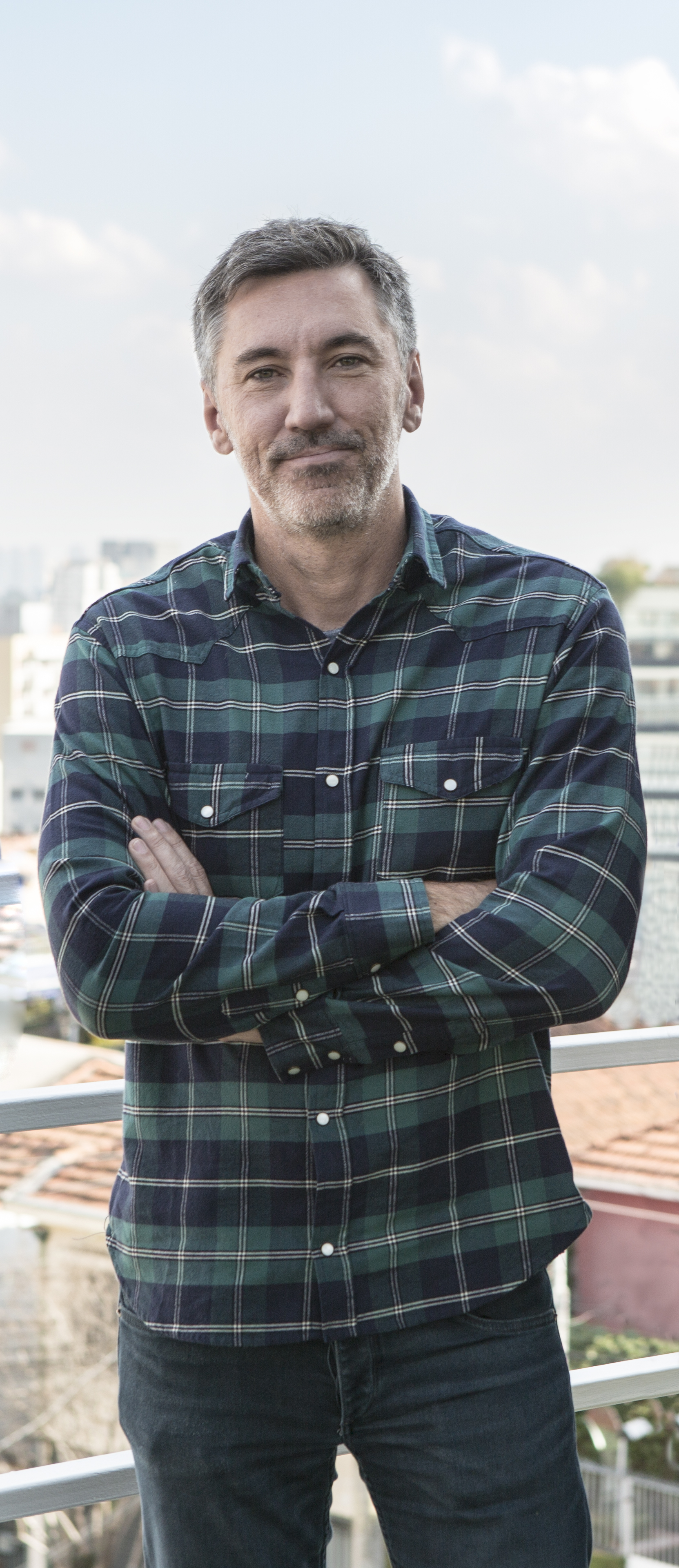 Homem branco grisalho posa de bra&ccedil;os cruzados vestindo camisa longa quadriculada nas cores preto e verde-escuro.