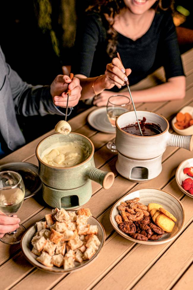 Confeitaria Dama Mesa de madeira com duas panelas de fondue, uma de queijo e outra de chocolate, ao lado de cumbucas com pão cortado e frutas.