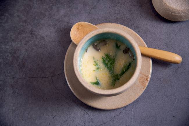 Cumbuca sobre prato em fundo cinza com creme amarelado ao lado de colher de madeira.