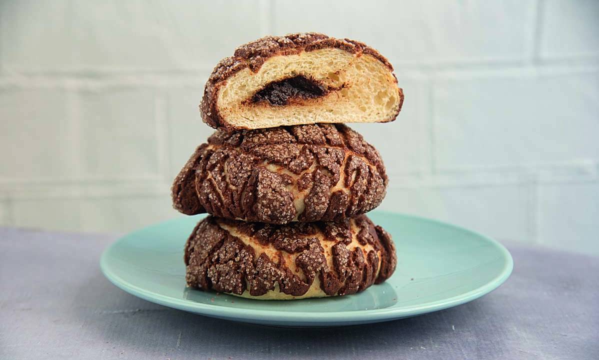 Três pãezinhos com casca de chocolate empilhados sobre prato de louça azul com o último aberto com recheio de chocolate.