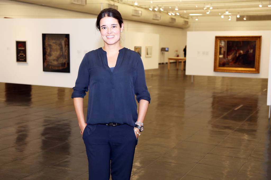 Juliana Sá__crédito_ MARINA MALHEIROS_DIVULGAÇÃO Mulher branca com cabelo preso posa com mãos no bolso em ambiente com quadros ao fundo. Veste roupa inteiramente azul.