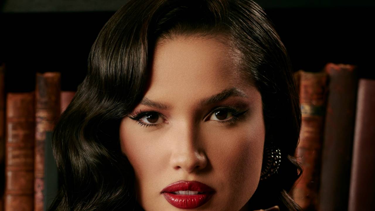 Mulher branca segura livro antigo com dedos indicadores em destaque. Tem cabelos pretos levemente encaracolados e batom bem vermelho.