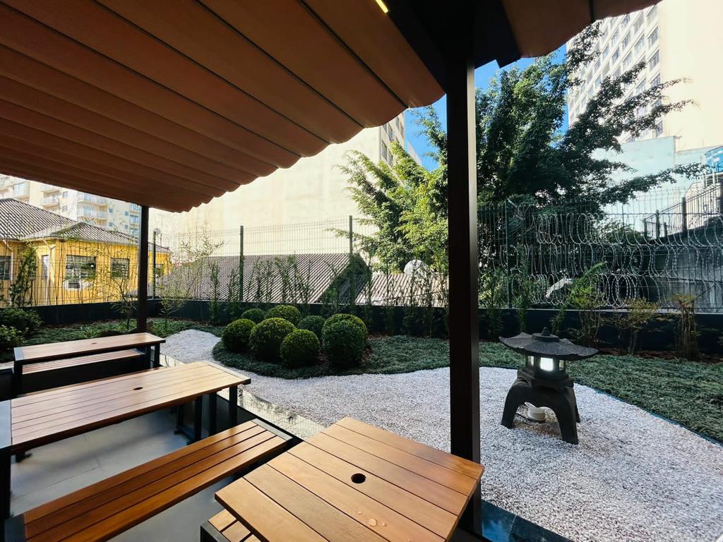 Liberdade 10 Do lado esquerdo, mesas e bancos de madeira, cobertos por um teto. Do lado direito, um pequeno jardim com caminhos de pedra e de grama. Uma árvore cerejeira ao fundo.