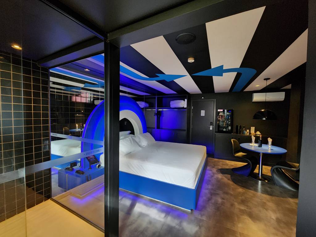 Suítes Andar de Cima-Crédito-Divulgação (1) Imagem de quarto de motel em tom azul e repleto de luzes neon, com cama de casal e chuveiro à esquerda. No teto, faixas em preto e branco com grandes setas azuis
