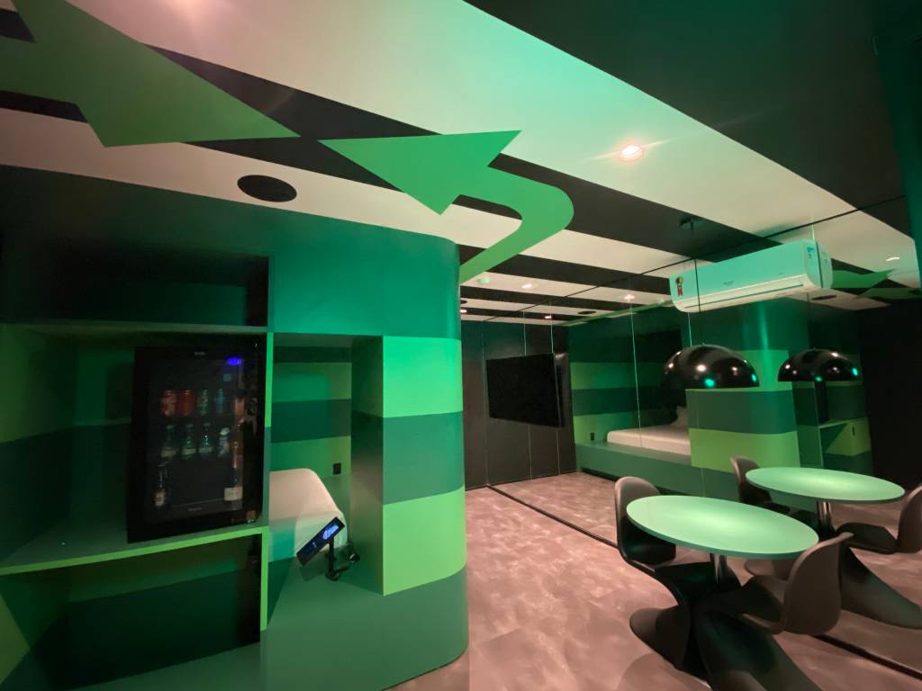 Suítes Andar de Cima-Crédito-Divulgação (10) Imagem de quarto de motel em tom verde e repleto de luzes neon, com cama de casal e chuveiro à esquerda. No teto, faixas em preto e branco com grandes setas verdes