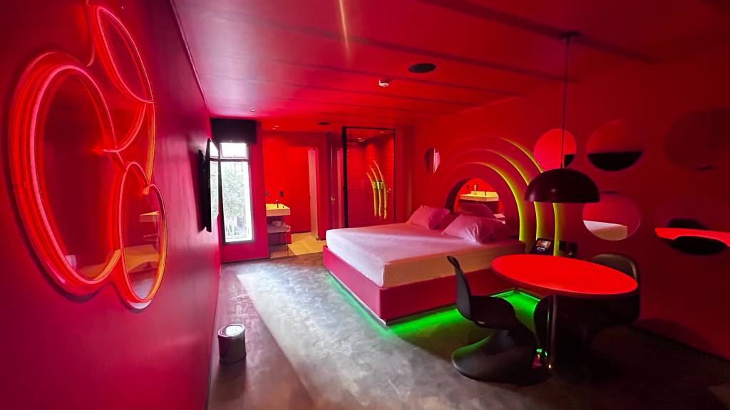 Suítes Andar de Cima-Crédito-Divulgação (2) Imagem de quarto de motel em tom vermelho e repleto de luzes neon, com cama de casal e chuveiro com janela ao fundo