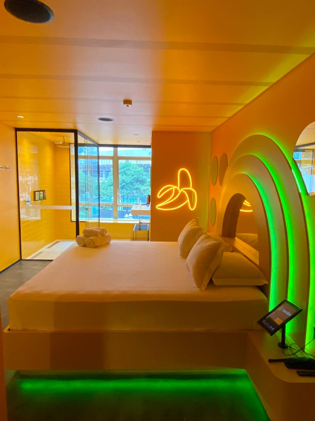 Suítes Andar de Cima-Crédito-Divulgação (3) Imagem de quarto de motel em tom amarelo e repleto de luzes neon, com cama de casal e chuveiro com janela ao fundo. Uma das paredes tem um neon em formato de banana.