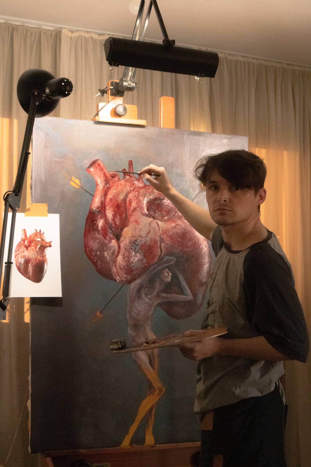 Susano Correia (Fran Bona-Divulgação) Moço branco de cabelos curtos castanhos posa em frente a um quadro com uma figura humana segurando uma espécie de coração gigante.