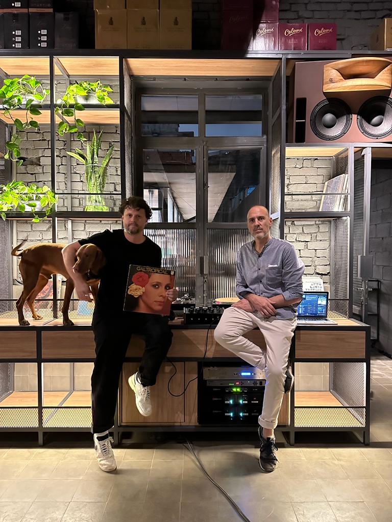 Elevado Conselheiro A imagem mostra os dois sócios do bar sentados em uma mesa com equipamentos de som. Leandro Mattiuz à esquerda segura um cachorro com um braço e com o outro um disco de Gal Costa.