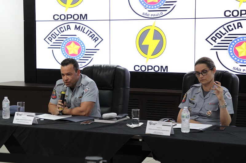 um policial e uma policial sentados à mesa. ele com um microfone.
