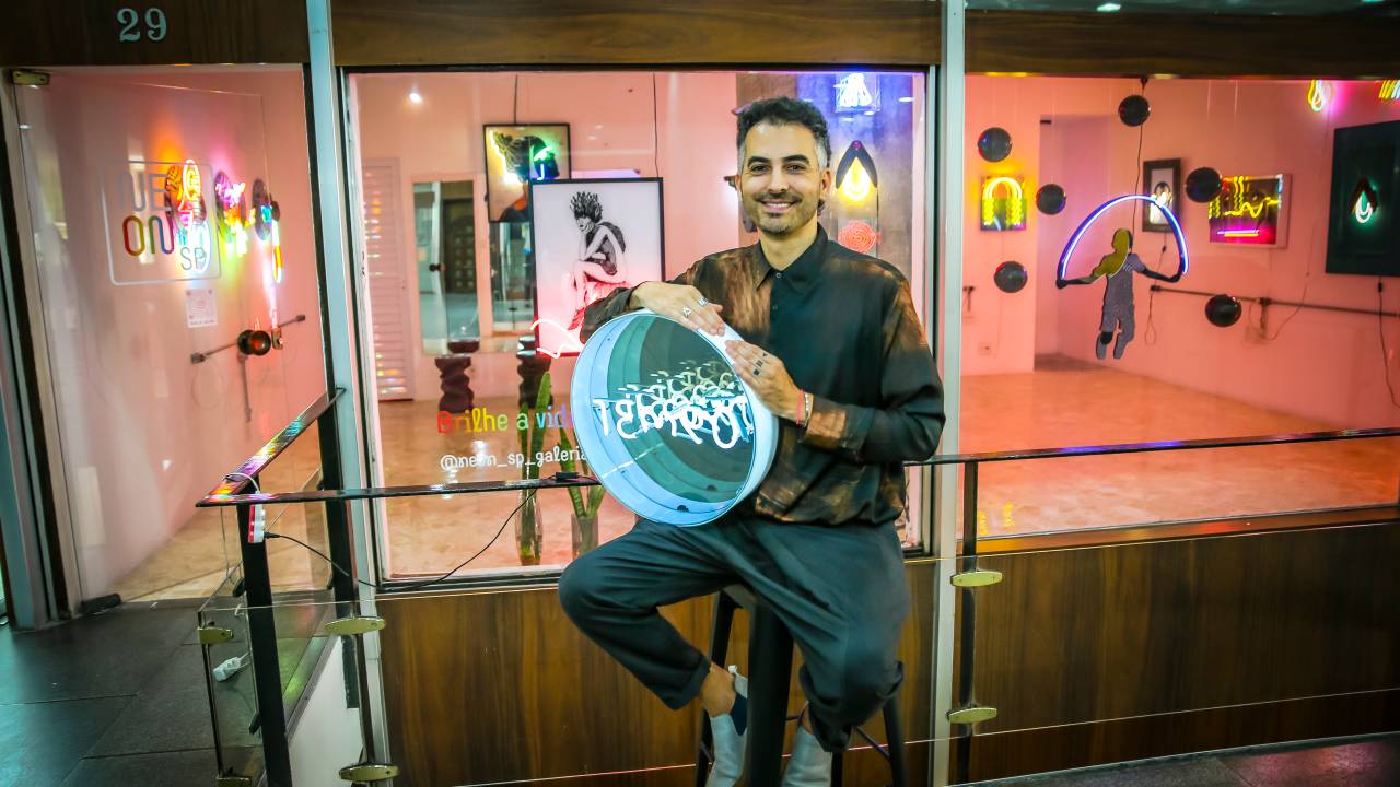 Homem branco levemente grisalho posa sentado segurando placa redonda neon. Veste roupas pretas e está em frente a sala repleta de luzes e quadros.