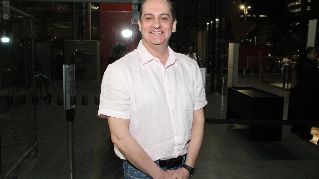 Homem branco de meia idade sorri segurando as mãos em frente ao corpo. Veste camisa branca curta e jeans.