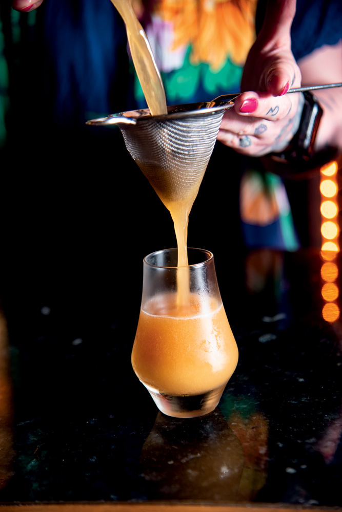 Bar dos Cravos A imagem mostra um copo de vidro com um drinque cor caramelo dentro. Uma pessoa despeja o drinque no copo com uma peneira.