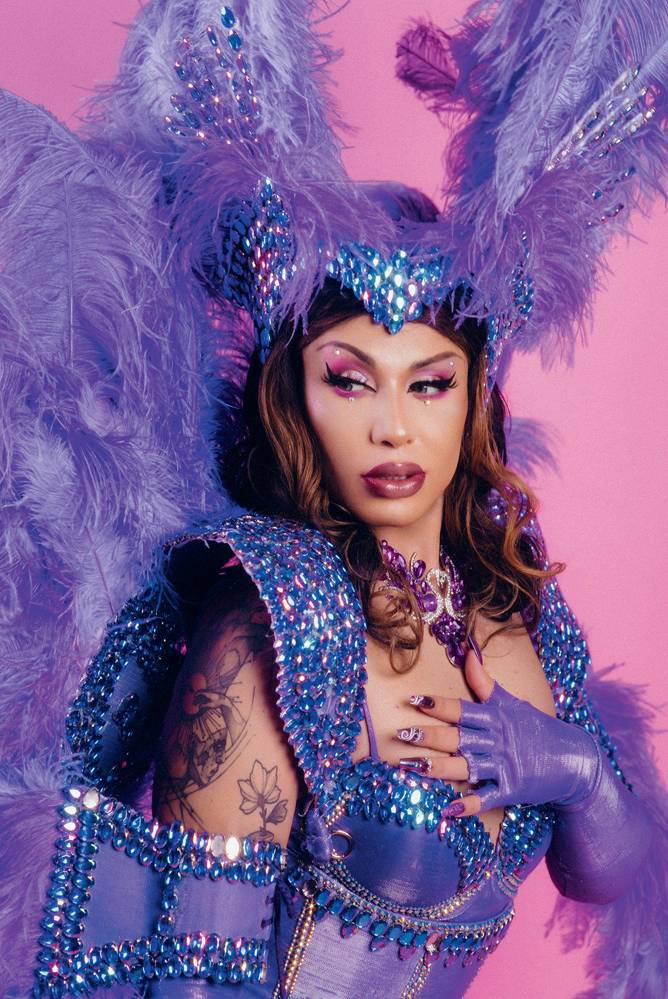 Grag Queen – Créditos Rodolfo Magalhaes (1).jpg Drag queen olha para o lado com uma mão no peito. Tem cabelos longos castanhos e veste adornos em tons roxos.
